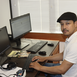 Ajay Shrestha sitting in his office, looking at the camera and in front of a keyboard and three computer screens