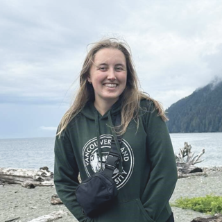 Emily Mahony outside in front of an ocean bay with mountains on a cloudy windy day smiling at the camera.