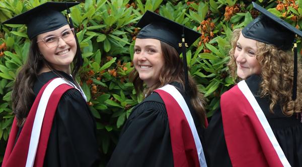 Three students wearing caps and gowns in front of a bush