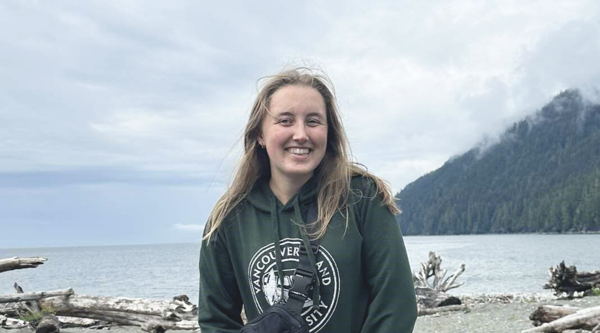 Emily Mahony outside in front of an ocean bay with mountains on a cloudy windy day smiling at the camera.