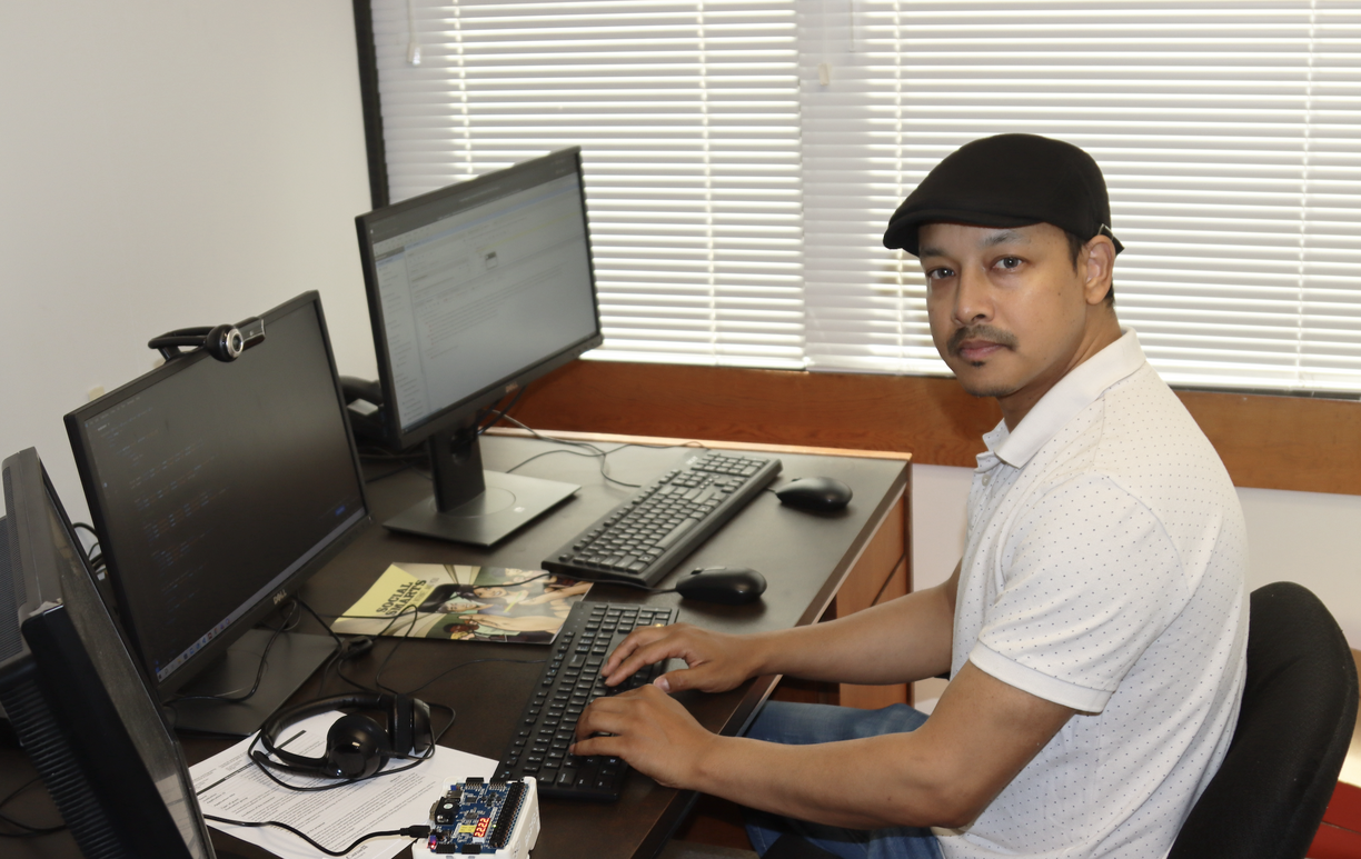 Ajay Shrestha sitting in his office, looking at the camera and in front of a keyboard and three computer screens
