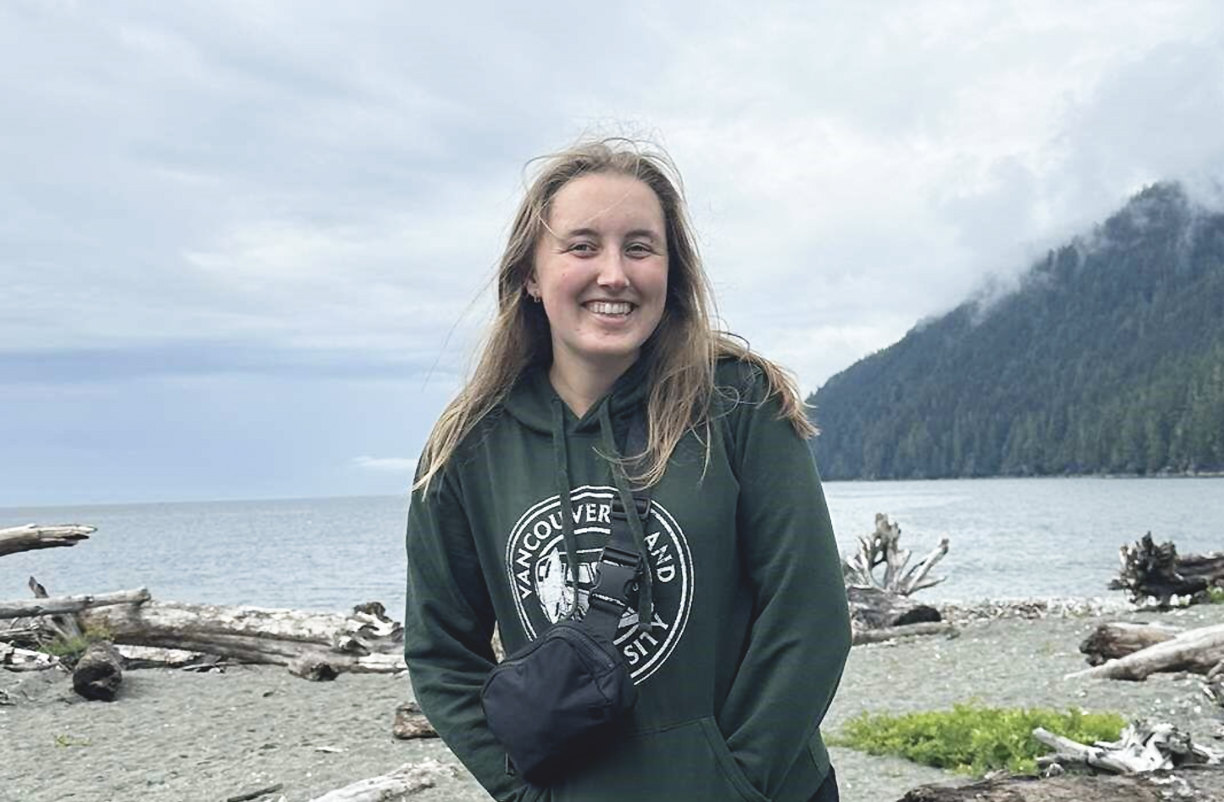 Emily Mahony outside in front of an ocean bay with mountains on a cloudy windy day smiling at the camera.