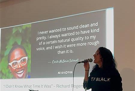 A Black woman speaks into a microphone with a PowerPoint slide behind her