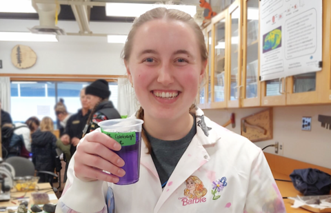 Emily Mahony in a chemistry lab, holding a cup of purple liquid and smiling at the camera.