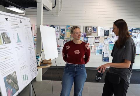Alysia Boudreau standing with a poster disply of her research at VIU and talking to another woman about it